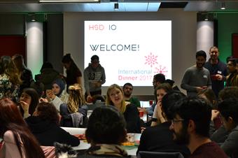 Viele internationale Studierende sitzen an Tischen und unterhalten sich. Im Hintergrund ist eine Präsentation an der Wand auf der steht Welcome International Dinner 2017.